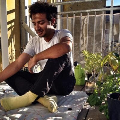 Full body photo of Zeerak, who is sitting on a white rug in a sunny outdoor patio, surrounded by potted plants. They have brown skin, short, wavy black hair, and are wearing a white t-shirt, black pants, and yellow socks.