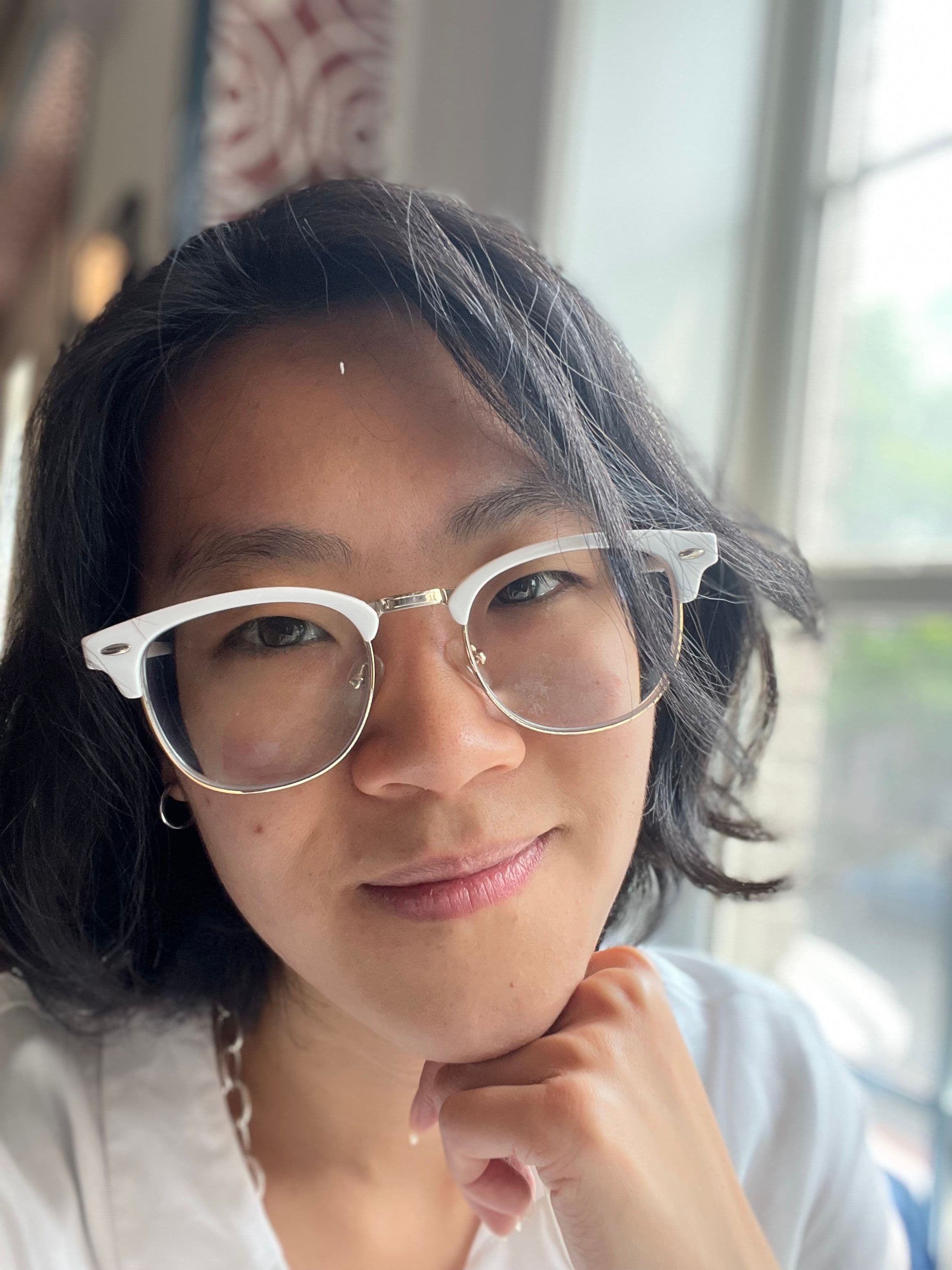 Headshot of Sophie song, who is Asian-American. They have straight, medium-length black hair, and are wearing white-framed glasses and a white shirt. They are smiling slightly, looking at the camera with their chin on one hand. Behind her is a window that seems to show a couple out of focus trees.