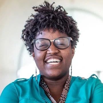 Photo of Nina, a Black woman with short, curly black hair. She is wearing glasses, long earrings, and a bright teal shirt. She has a big smile. The background of the photo is out of focus and white.