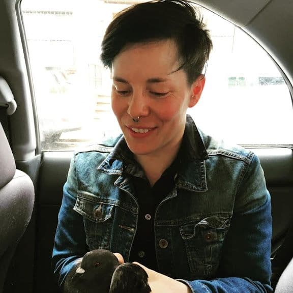 Photo of Christie, a white person with short brown hair. They appear to be sitting in a car, and holding a pigeon in their hands. She is wearing a denim jacket, and has a silver-colored ring in her nose. She is smiling down at the bird.